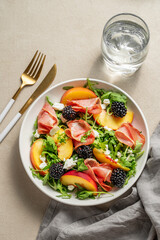 Fresh salad with peaches, arugula, feta, prosciutto and berries in a plate on a light background