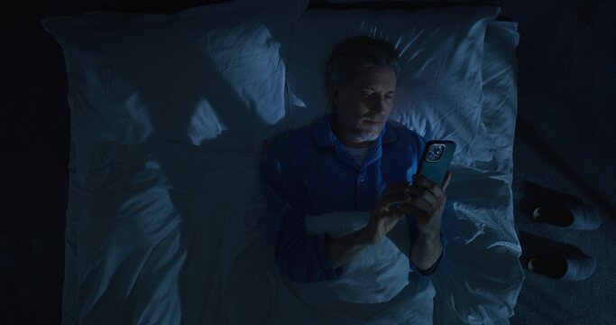 Mature Man Lies in White Bed Under Blanket, Using Smartphone Before Sleep. Concept of Screen Addiction, Poor Sleep Hygiene, Late Night Scrolling, Insomnia, Modern Lifestyle Habits. Static Shot.