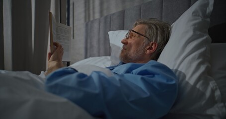 Waking Up in Morning in Bright, Cozy Bedroom, Mature Man Wearing Glasses Lies Relaxed in White Bed Reading Book. Pleasant, Leisurely Morning, Relaxing in Bed. Concept of Morning Routine. Static Shot.