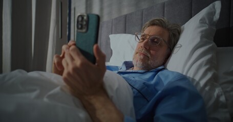 Fototapeta premium In Morning, in Sunny Bedroom, Man Wearing Glasses Lies in Bed With Smartphone, Intently Reading News. Morning Routine, Checking Messages, News on Internet, Social Networks. Concept of Digital Habits.