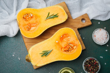 Butternut squash halved with rosemary sprigs on wooden board with salt and peppercorns