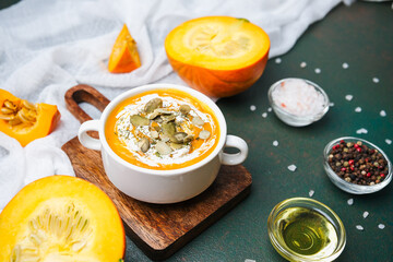 Bowl of creamy pumpkin soup topped with seeds, served with whole pumpkin and spices on a dark surface.