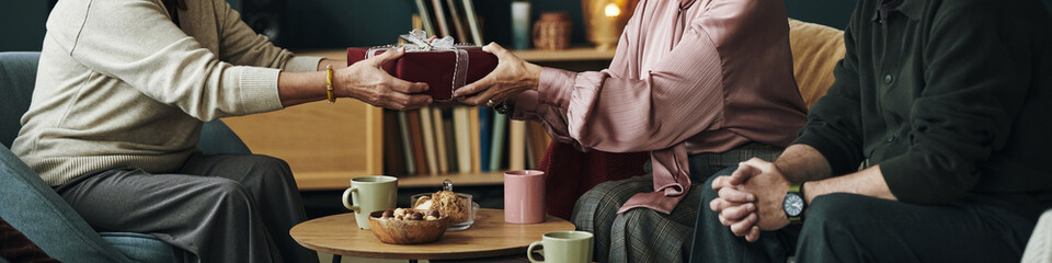 Header of senior Caucasian woman giving wrapped gift to female friend while male companion sitting nearby, three people gathering around coffee table with mugs