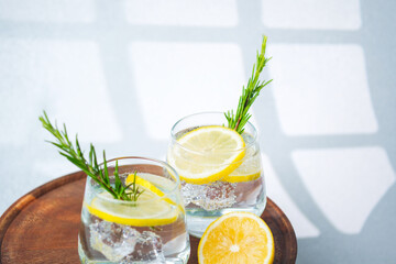 Lemon water with rosemary sprigs and ice cubes served in glasses on round wooden table beside fresh citrus halves
