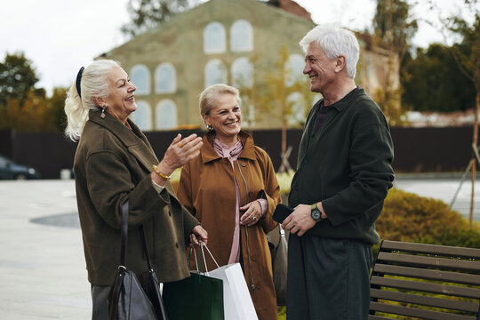 Group of two senior Caucasian women and one man standing outdoors smiling and talking, holding shopping bags, engaging in friendly conversation near bench in urban setting - Powered by Adobe