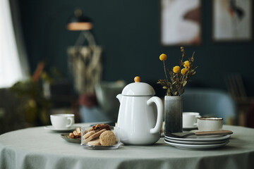 Ceramic teapot standing on round table with stacked plates, cups, cookies on glass tray, dried flower bouquet in vase, blurred modern living room interior in background