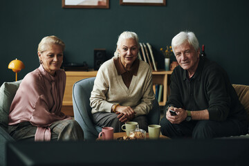 Portrait of three senior Caucasian people sitting together on sofa watching television, two women and one man smiling and looking toward camera, coffee mugs on table