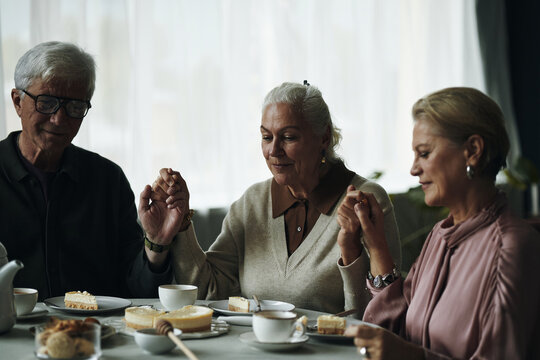 Senior Caucasian man and two elderly women friends sitting at table holding hands before eating dessert, engaging in moment of gratitude or prayer, sharing time together indoors