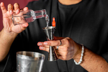 Bartender pouring clear liquid into a jigger, preparing a cocktail. Close-up of hands with bracelet and ring. Professional mixology scene with blurred bar background.
