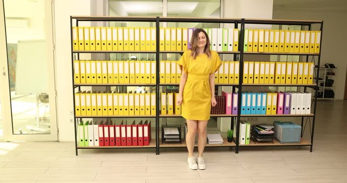 Pretty woman employee in yellow dress dances near ring binders rack in office. Positive female clerk expresses joy and happiness at workplace