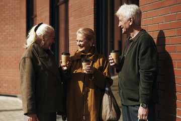 Three senior Caucasian people standing outdoors near brick wall, holding takeaway coffee cups, smiling and talking, two women and one man engaging in friendly conversation