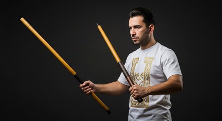 A man wielding two wooden sticks dressed in a white tshirt against a dark background