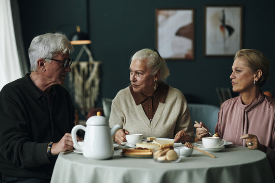 Three senior Caucasian people sitting at round table eating dessert and drinking coffee, two women and one man engaging in conversation, pastries and teapot on table during friendly gathering - Powered by Adobe