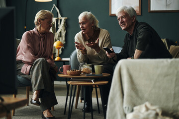 Three senior Caucasian friends sitting together, engaging in animated conversation while watching television, two women and one man smiling and gesturing around coffee table