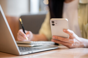 Close up woman student holding pen writing in notebook looking at phone aside laptop on table in cafe