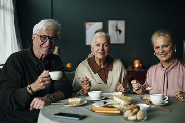 Three senior Caucasian people sitting at table enjoying dessert, with tea together, smiling and looking at camera, holding cups and plates with pastries and bread, sharing friendly moment