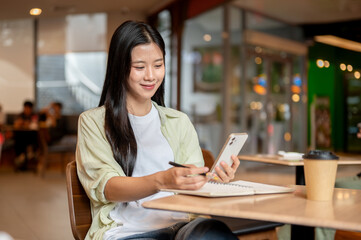 Fototapeta premium Asian woman student looking at phone holding pen over notebook aside coffee at wooden table in cafe.
