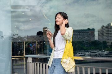 Asian woman holding phone carrying yellow bag brushing hair back leaning on shopping mall's railing.