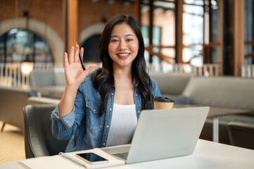 Fototapeta premium Asian woman student holding coffee waving hand with pen in front of laptop and book on table in cafe
