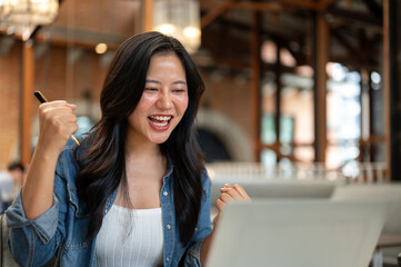 Asian woman holding pen and making a fist in excited in front of a laptop while sitting in the cafe.