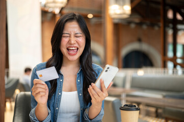 Asian woman holding credit card and phone while screaming in excited at table in cafe or coffee shop