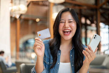 Asian woman holding credit card and phone while laughing in excited at table in cafe or coffee shop