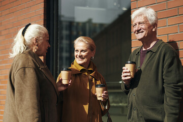 Three Caucasian seniors standing outdoors holding takeaway coffee cups, two women and one man smiling and talking together in front of brick wall on city street