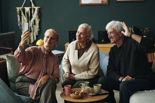 Fototapeta Three Caucasian seniors sitting together, smiling and posing for selfie with smartphone, older man waving hand while two senior women looking at camera, drinks and snacks on table