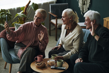 Three senior Caucasian people sitting together waving at smartphone during video call with friends, two women and one man smiling and interacting, coffee table with snacks and cups in foreground