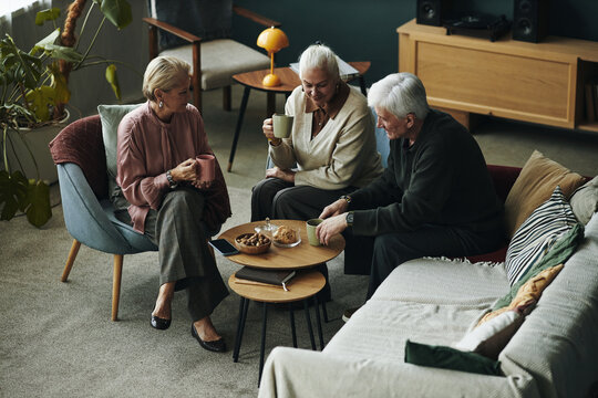 Fototapeta Three senior Caucasian women and man sitting together in living room drinking hot beverages and talking, snacks, cookies and biscuits on coffee table, relaxed social gathering among friends