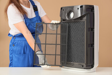 Female technician changing dirty air purifier filter on beige background
