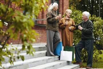 Three senior Caucasian people standing on outdoor steps holding takeaway coffee cups, engaging in...