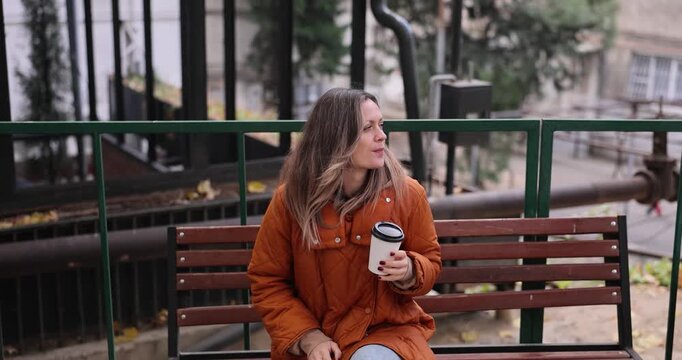 Woman sits comfortably on bench and enjoys warm coffee in autumn. Female gazes thoughtfully at busy city square and sips slowly from paper cup