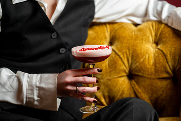 Elegant woman holding pink cocktail in coupe glass, seated on mustard-yellow tufted sofa in upscale lounge. Stylish attire, dark nails, and red garnish add sophistication.