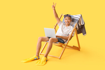 Young Asian man with laptop and flippers showing victory gesture on deckchair against yellow background © Pixel-Shot
