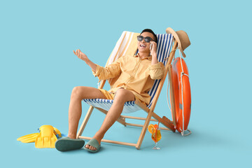 Young Asian man with mobile phone and beach accessories sitting on deckchair against blue background