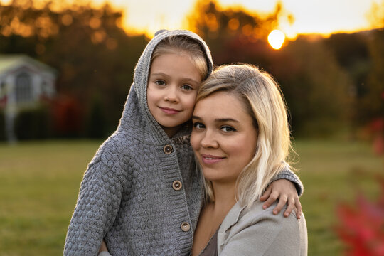 Happy family mother and little daughter look at camera and smile on background of autumn park at sunset. Weekend leisure walking with child outdoors