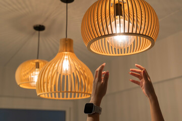 Hand reaching to adjust modern wooden pendant ceiling lamp with glowing light bulb interior home decoration