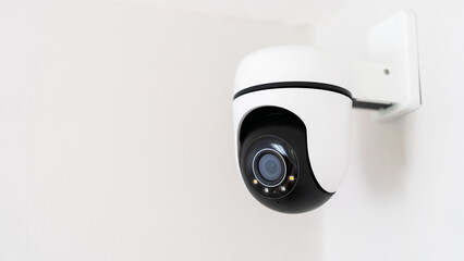 A camera is mounted on a wall. The camera is black and white. The camera is pointed at the viewer