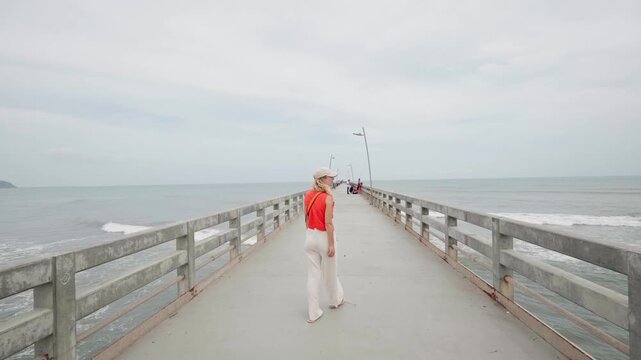 Young woman walking away from the camera along a long pier towards the sea