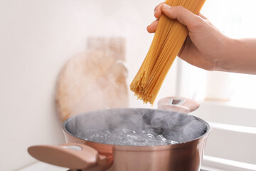 Woman putting spaghetti into cooking pot with boiling water on electric stove in kitchen