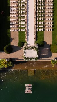 drone view of luxury outdoor wedding ceremony on Lake Como with floral arch, rows of white chairs on lush green lawn, and drone flying back to reveal panoramic lake and mountain views
