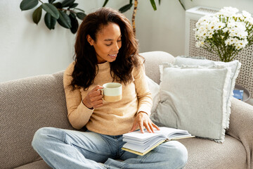 African American woman relaxing reading book on sofa drinking coffee