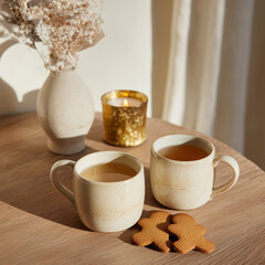 Two ceramic cups of tea on a small wooden table with gingerbread cookies, a tiny golden candle and a mini vase with