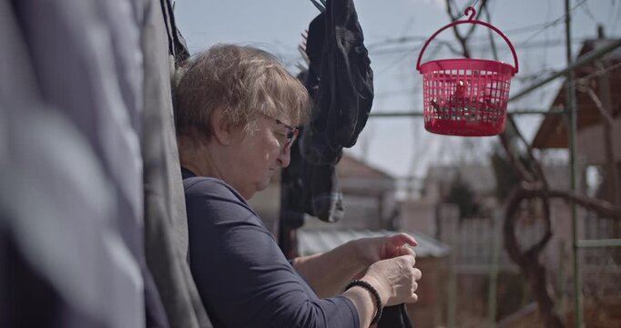 An elderly woman hangs laundry on a clothesline in her backyard. She uses clothespins to secure the items, which include socks and other garments.