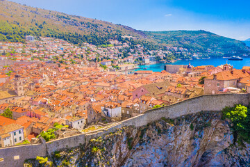 Aerial scenic view of Dubrovnik old town showing ancient city walls rising above the rooftops, blue Adriatic sea, historic architecture and marble Mediterranean cityscape