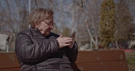 An elderly woman enjoys a sunny day in the park while sitting on a bench and using her mobile phone. - Powered by Adobe
