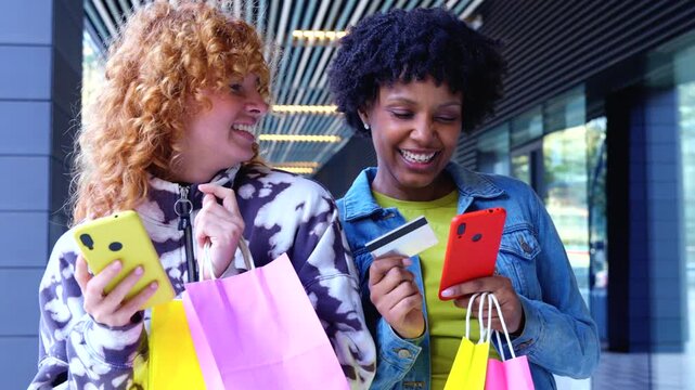 Cheerful African American and Caucasian friends stand in a mall atrium holding colorful bags, browsing a yellow smartphone,one taps a credit card,illustrating cashless online shopping ease rightnow 