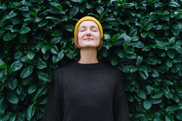 Portrait of relaxed and confident woman with eyes closed on background of green leaves wall. Thoughtful person in front of green hedge. Stability through mental health. Self-care, life balance concept