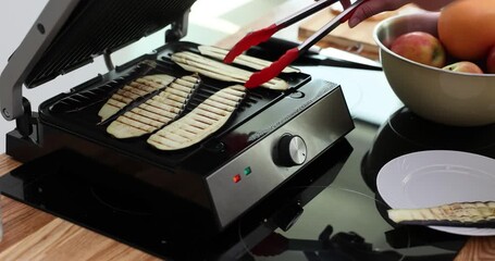 Woman hand with tongs turns over sliced eggplants on electric grill at home. Female cooks tasty zucchini with kitchen device indoors slow motion